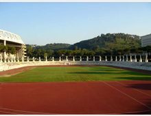 Vista di uno stadio con pista di atletica rossa e campo erboso centrale, circondato da gradinate basse e una fila di statue lungo il bordo. Sullo sfondo si vedono colline alberate e alcuni edifici ai lati.