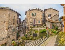 Borgo storico in pietra con case ravvicinate e facciate in muratura, finestre con scuri e tetti in coppi. In primo piano una scalinata e una strada in pendenza con gradini, piccoli muretti e aiuole, tra vicoli stretti e costruzioni addossate.