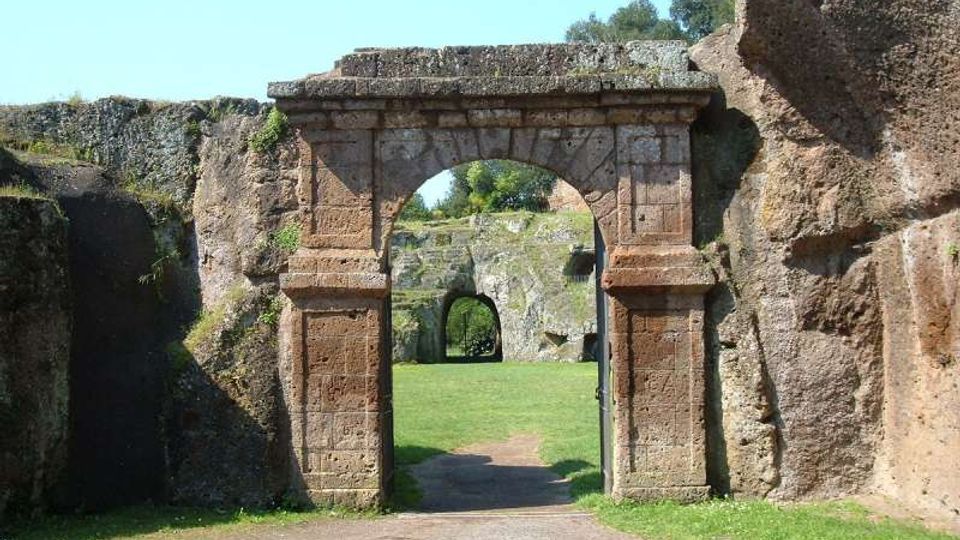 Portale in pietra con arco centrale incastonato tra alte pareti rocciose, che introduce a un prato con un sentiero. In fondo si vede un secondo arco più piccolo ricavato nella roccia.