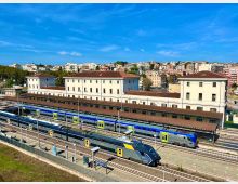 Vista panoramica di una stazione ferroviaria con più binari e treni blu in sosta sotto pensiline lunghe. Sullo sfondo si vede un grande edificio storico chiaro con tetti in tegole rosse e, oltre, un quartiere collinare con palazzi.
