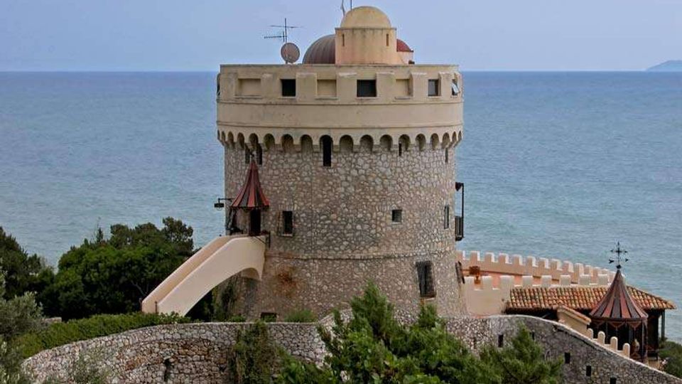 Torre costiera in pietra di forma cilindrica con merlature e terrazza superiore, collegata da una rampa a un complesso con mura e piccoli tetti in tegole. Sullo sfondo si vede il mare, con vegetazione bassa e alberi in primo piano.