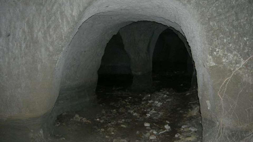 Ingresso ad arco di una grotta o galleria sotterranea con pareti in roccia grezza. Il pavimento è irregolare, coperto di sassi e detriti, e l’interno si apre in più passaggi bui.