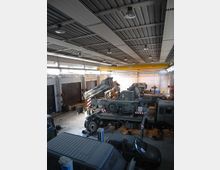 Interno di un grande capannone industriale adibito a officina, con tetto in lamiera e illuminazione a sospensione. Sul pavimento sono presenti vari veicoli militari e camion in manutenzione, attrezzature da lavoro e un carroponte giallo che attraversa la struttura.