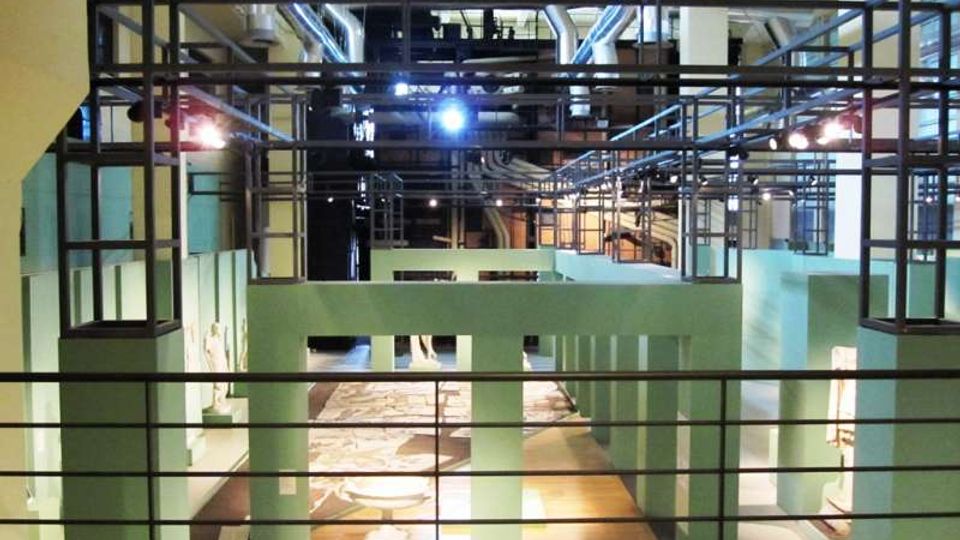 Interno di una sala espositiva in stile industriale, con travi e tubature a vista sul soffitto e una struttura metallica sospesa. Pareti verde acqua delimitano aree con reperti e pavimenti decorati, osservabili da una balconata con ringhiera in primo piano.