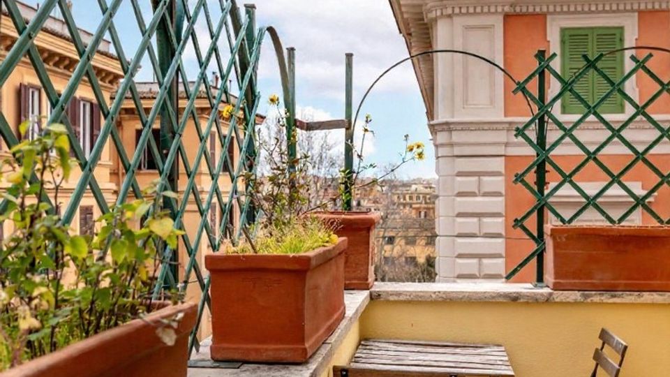 Terrazza urbana con muretto perimetrale e grigliati verdi, fioriere rettangolari in terracotta e un piccolo tavolo con sedia pieghevoli in legno. Sullo sfondo si vedono facciate di edifici con persiane verdi e cielo parzialmente nuvoloso.