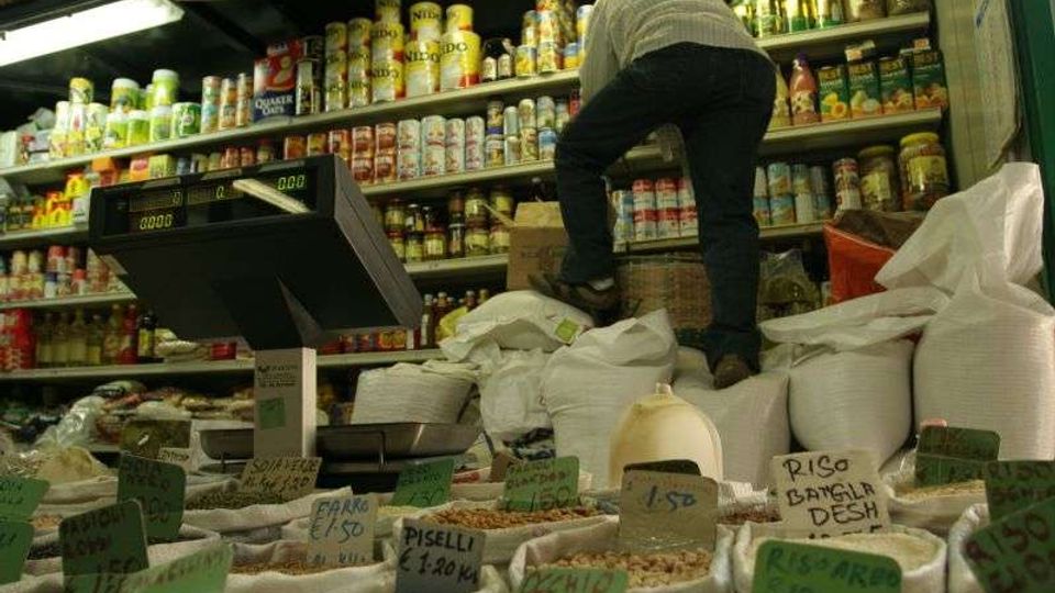 Interno di un negozio di alimentari con sacchi aperti di cereali e legumi in primo piano, etichettati con cartellini di prezzo. Sullo sfondo ci sono scaffali alti pieni di conserve e prodotti confezionati, con una bilancia digitale sul bancone.