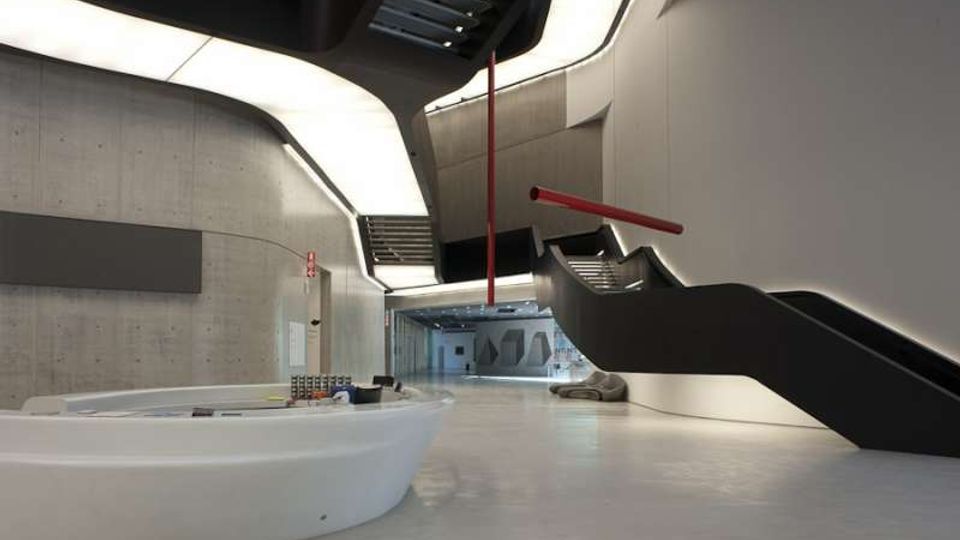 Interno di un edificio moderno con ampio atrio in cemento e superfici bianche, illuminato da luci integrate nel soffitto curvo. Due scale nere con corrimano rossi salgono ai piani superiori e al centro si trova un banco reception ovale bianco su pavimento lucido.