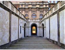 Passaggio pedonale in pietra tra alte pareti intonacate, che conduce a un portico con arcate. In fondo si vede la facciata di un edificio storico con finestre ad arco, colonne e balconata sopra un grande arco centrale.
