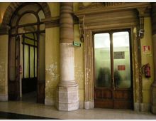 Ingresso interno di un edificio storico con pareti gialle consumate, colonne in pietra e cornici decorative attorno a porte in legno con vetri opachi. A sinistra c’è un grande arco vetrato che conduce a un corridoio, mentre a destra una porta con insegna “U.R.P.” è affiancata da segnaletica e un estintore a parete.
