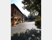 Piazzale esterno con pavimentazione in sampietrini, affiancato da un edificio a due piani color ocra con cornici rosse e finestre ad arco. Sullo sfondo si vedono alberi alti e un’area di parcheggio, con rami che proiettano ombre sull’acciottolato.