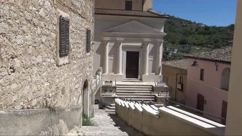 Scalinata in pietra tra edifici in muratura che conduce a una chiesa con facciata chiara, frontone triangolare e portico con colonne. Sullo sfondo si vedono case del borgo e colline verdi sotto un cielo sereno.