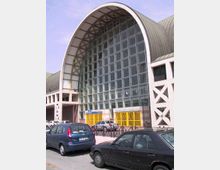 Facciata di un grande edificio pubblico con ampia vetrata a griglia e copertura ad arco in metallo, affiancata da strutture laterali con elementi a X. Davanti si trovano cancelli gialli d’ingresso e un’area stradale con auto parcheggiate.
