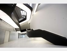 Atrio interno moderno e minimalista con ampie pareti bianche e in cemento e pavimento chiaro. Una scala nera scultorea con linee curve sale verso un lucernario superiore, con passerelle e aperture su più livelli.