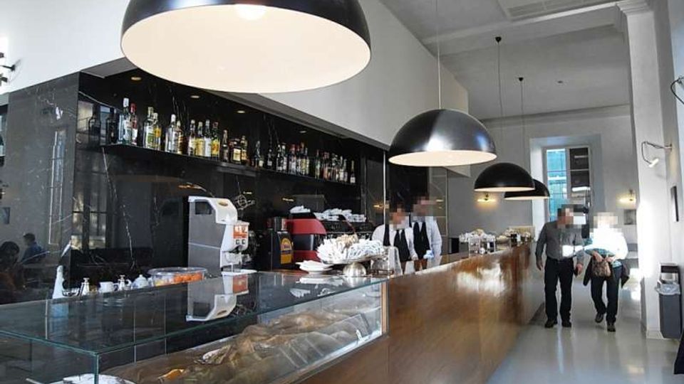Interno di un bar moderno con lungo bancone in legno e vetrina espositiva, retrobanco nero con scaffali di bottiglie e attrezzature. Grandi lampade a sospensione a cupola illuminano lo spazio con pareti bianche e soffitto alto.