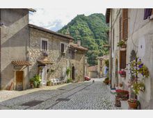 Strada acciottolata in un borgo collinare, fiancheggiata da case in pietra e intonaco con persiane in legno, archi e vasi di fiori sui davanzali. Sullo sfondo si vedono un campanile e una collina boscosa che domina l’abitato.