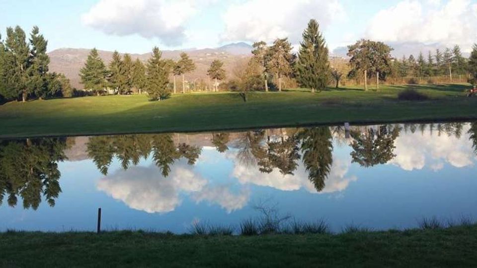 Campo verde alberato con un lago in primo piano che riflette alberi e nuvole; sullo sfondo si vedono colline e montagne sotto un cielo parzialmente nuvoloso.