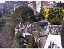 Terrazza panoramica in pietra su un pendio, con muretti, scale e ringhiere che collegano piccoli terrazzamenti con alberi e piante in vaso. Sullo sfondo si vedono case addossate tra loro lungo il bordo del borgo e la valle sottostante.