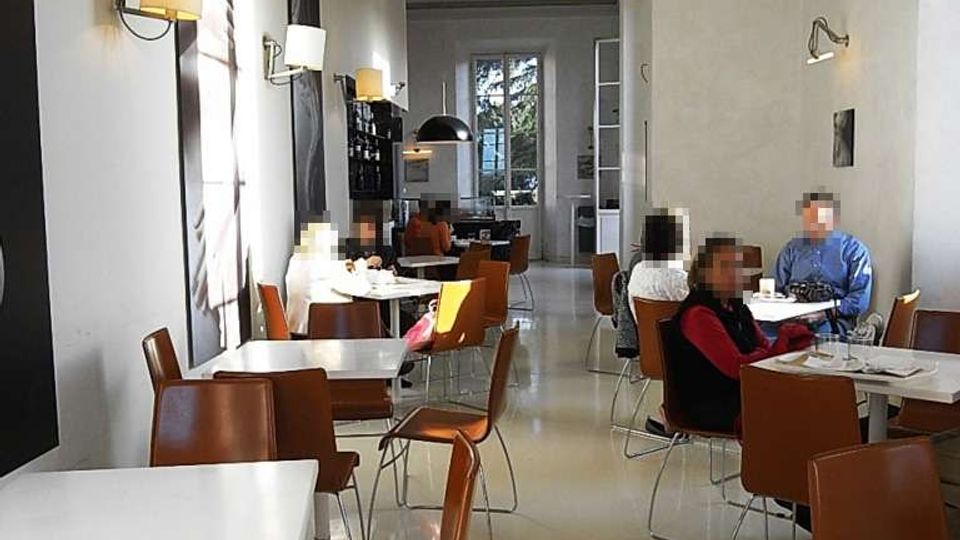 Interno di una caffetteria moderna e luminosa con pareti bianche alte, grandi stampe a parete e applique. Tavoli quadrati bianchi con sedie marroni sono disposti lungo una sala stretta, con una grande finestra in fondo che lascia entrare luce naturale.