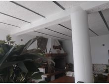 Interno di un soggiorno con pareti bianche e soffitto a travi, con una grande colonna cilindrica in primo piano. Sullo sfondo si vede un camino in pietra con mensola in legno e arredi da salotto; una pianta con foglie ampie occupa parte della vista a sinistra.