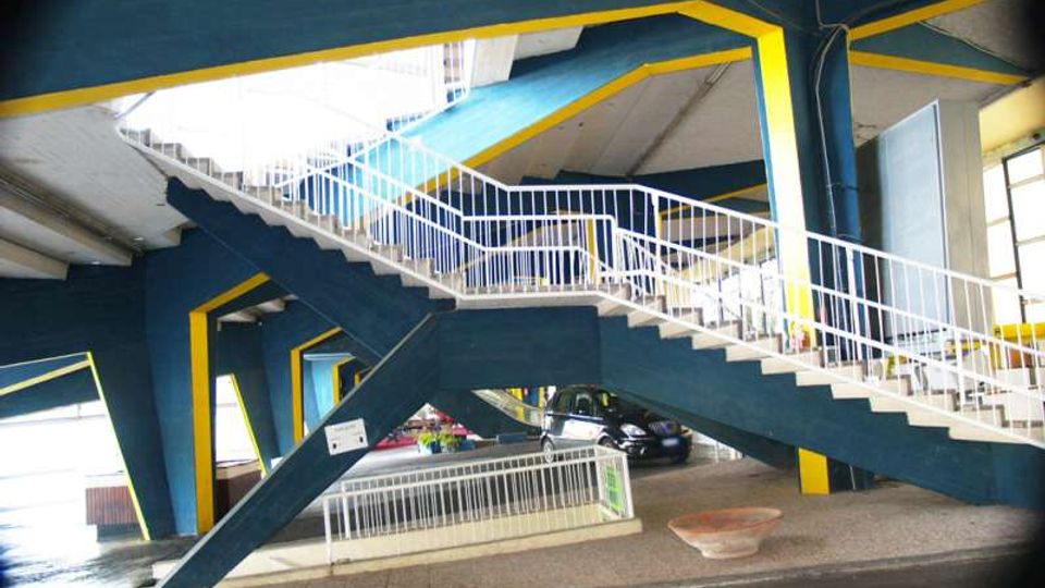 Interno di una struttura in cemento con scala pedonale sopraelevata a rampe incrociate, ringhiere bianche e travi blu con bordi gialli. Sotto la scala si vede un’area di passaggio coperta con pavimentazione e una corsia carrabile.