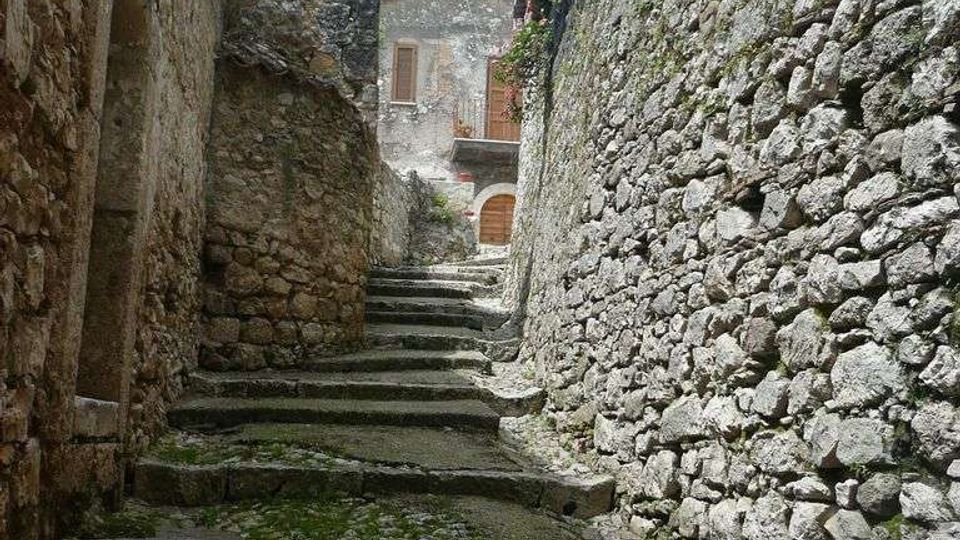 Vicolo in pietra in salita con gradini irregolari e pavimentazione acciottolata, fiancheggiato da alti muri in muratura grezza. In fondo si vede la facciata di una casa con porta ad arco e finestre, con vegetazione che sporge dal muro a destra.