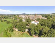 Veduta aerea di un borgo con edifici in pietra e una torre campanaria, circondato da ampie aree verdi e alberate. In primo piano si vede un grande edificio chiaro con tetto in coppi vicino al margine di una scarpata e a campi aperti.