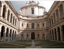 Cortile interno di un complesso storico con porticati ad archi su due livelli lungo i lati e, sul fondo, una facciata curvilinea con finestre e grande portale, sormontata da una cupola. Pavimentazione con disegni geometrici in pietra e aree verdi.