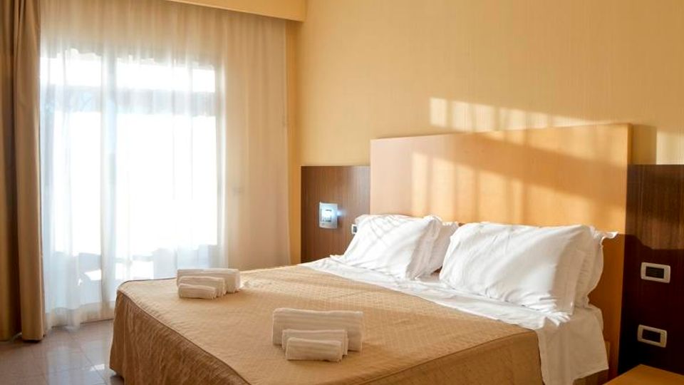 Camera da letto d’albergo con letto matrimoniale, testiera in legno e comodino, pavimento piastrellato e pareti color beige. Una grande finestra con tende lascia entrare luce naturale.
