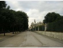 Lungo viale alberato con strada sterrata che conduce a un edificio monumentale sullo sfondo, con facciata classica decorata da statue e una cupola centrale. A destra corre un alto muro con ringhiera in ferro, mentre a sinistra ci sono alberi e lampioni lungo il margine del percorso.