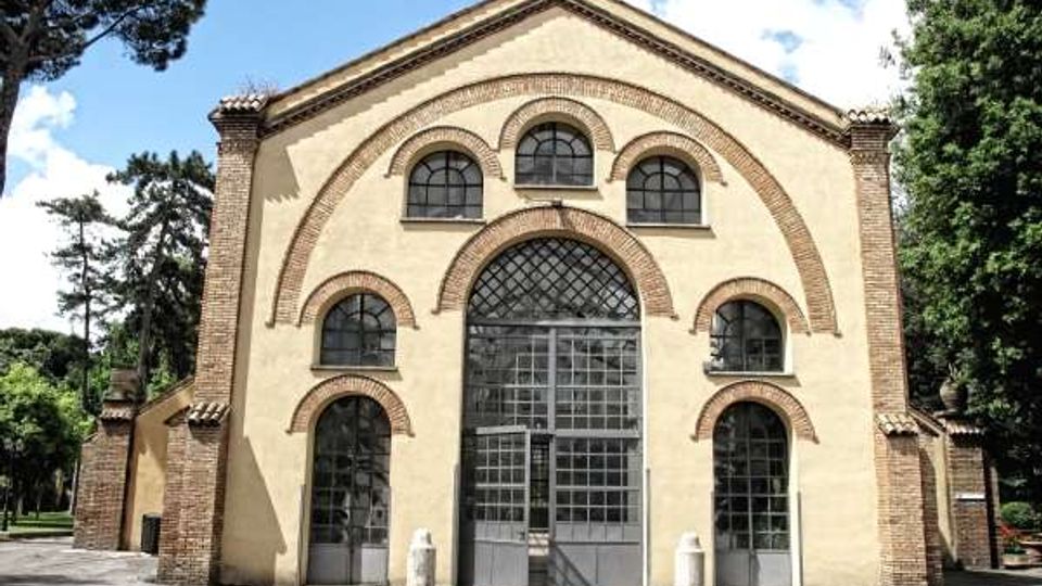 Facciata di un edificio in stile romanico con intonaco chiaro e cornici in mattoni, caratterizzata da grandi archi sovrapposti e ampie finestre ad arco con vetri a griglia. L’ingresso centrale è più alto e vetrato, e l’edificio è circondato da alberi e da un piazzale asfaltato.
