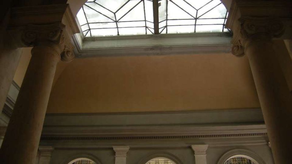 Interno di un edificio storico con grandi colonne e arcate inferiori, illuminato da un lucernario a volta con vetrata geometrica. Pareti intonacate e cornici decorative incorniciano l’atrio a doppia altezza.