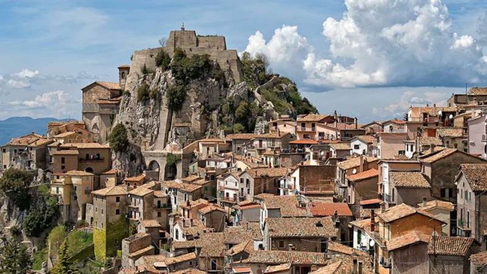 Veduta panoramica di un borgo storico con case addossate e tetti in tegole, disposto su un pendio roccioso. In alto domina una grande fortificazione in pietra costruita sulla rupe, con cielo parzialmente nuvoloso sullo sfondo.