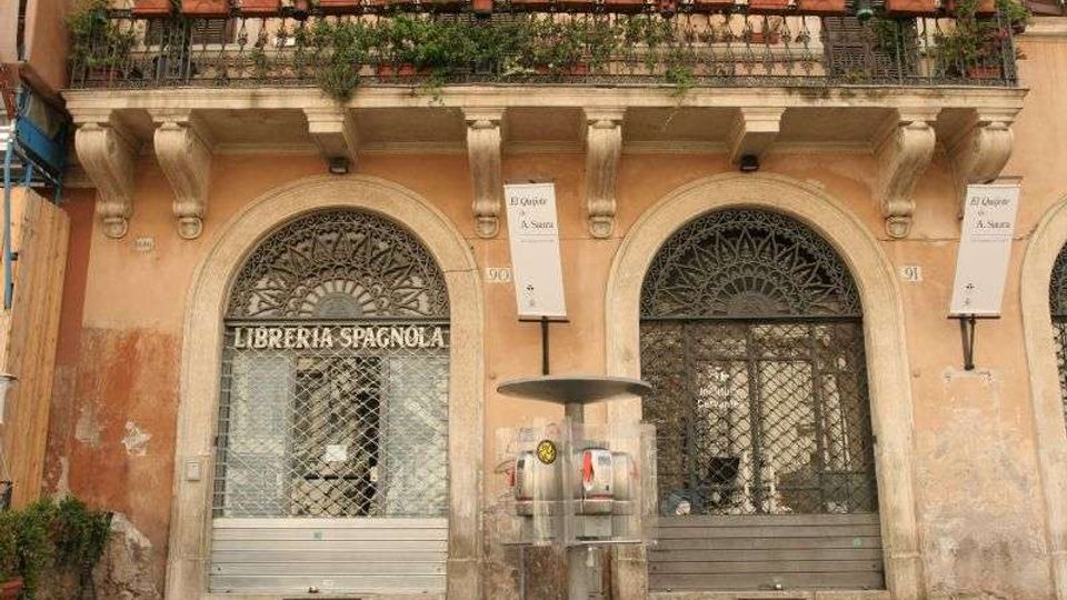 Facciata di un edificio storico con due grandi archi al piano terra chiusi da serrande e grate decorative, e una balconata superiore con ringhiera in ferro e molte piante in vaso. Al centro si vede una piccola fontanella su un marciapiede in pietra.