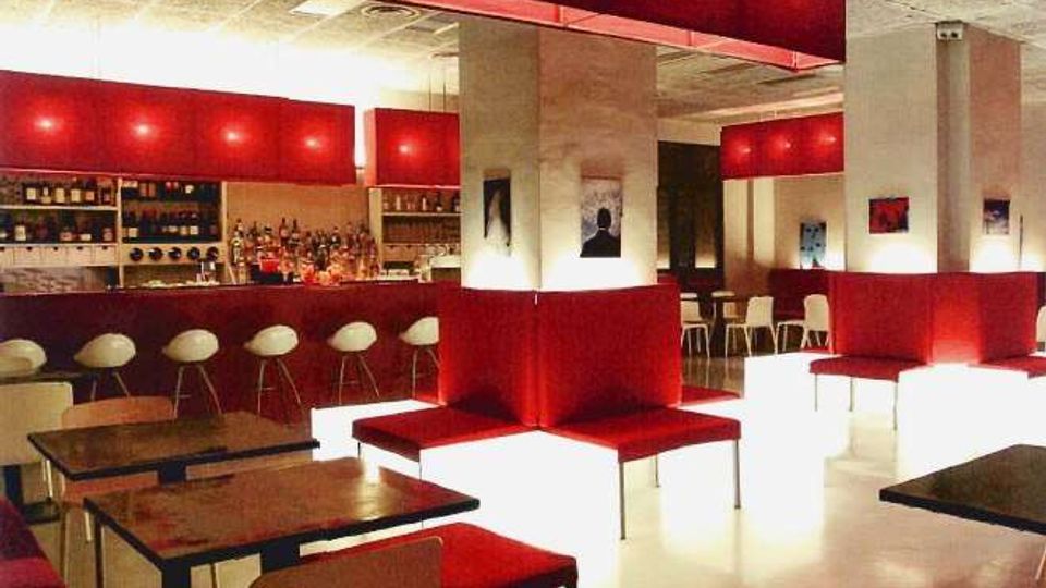 Interno di un bar moderno con arredi rossi e bianchi, bancone illuminato e mensole con bottiglie sullo sfondo. Sedute imbottite a panche, tavolini e sgabelli bianchi sono disposti in una sala ampia con colonne e grandi lampade rettangolari sospese.