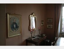 Interno di una stanza in stile classico con pareti marroni, un tavolo consolle in legno con candelabri e oggetti decorativi, e uno specchio con cornice dorata sopra. Sulle pareti sono appesi un ritratto incorniciato e alcune stampe, accanto a una finestra con tende pesanti.