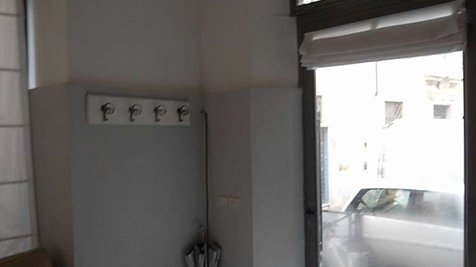 Interno di un piccolo locale con pavimento in graniglia, pareti bianche e una grande porta/vetrina d’ingresso con sopraluce decorato in ferro battuto. Sono visibili una lampada a sospensione, un pannello appendiabiti a parete e una finestra laterale.