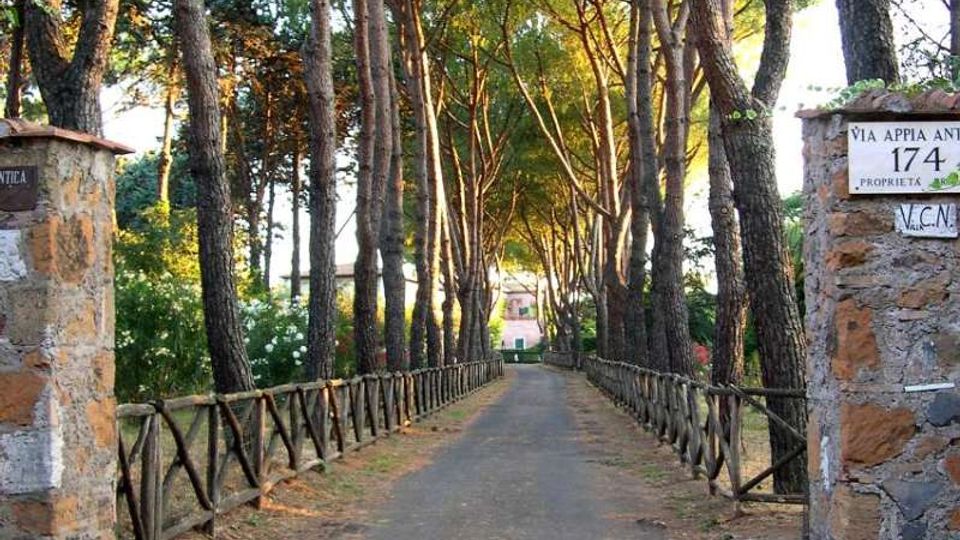 Viale asfaltato rettilineo fiancheggiato da alti pini e da staccionate in legno, che conduce verso edifici sullo sfondo. All’ingresso ci sono pilastri in pietra con targa “Via Appia Antica 174”.
