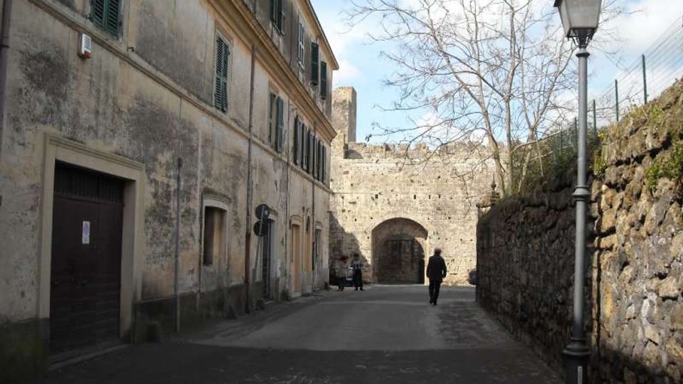 Strada lastricata in un borgo storico, affiancata da un lungo edificio intonacato con persiane verdi e da un alto muro in pietra con lampione. Sullo sfondo si vede una porta ad arco inserita in una fortificazione in pietra.