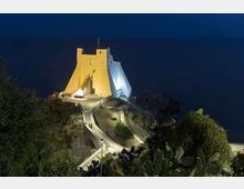 Edificio bianco dalle forme moderne e angolari, illuminato di notte, su un promontorio roccioso affacciato sul mare. Una strada tortuosa e una scalinata conducono all’ingresso, circondate da vegetazione mediterranea.