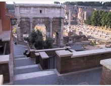 Veduta dall’alto di un arco monumentale romano in pietra con tre fornici e iscrizioni, affacciato su un’area archeologica con resti di edifici e colonne. In primo piano si vede una terrazza con muretti in mattoni e una scalinata pavimentata.