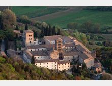 Veduta aerea di un complesso monastico in pietra con vari edifici a tetti in tegole e un campanile centrale, disposto attorno a cortili interni. Il monastero è circondato da alberi e colline con campi coltivati sullo sfondo.