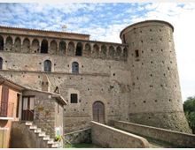 Facciata in pietra di un castello medievale con torre cilindrica merlata e loggiato ad arcate lungo il livello superiore. In primo piano si vedono una rampa murata e una piccola costruzione addossata alle mura, sotto un cielo parzialmente nuvoloso.