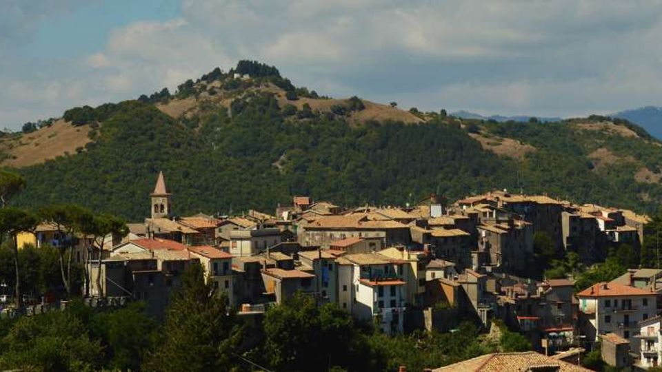 Veduta panoramica di un borgo collinare con case ravvicinate dai tetti in tegole e un campanile che svetta tra gli edifici. Sullo sfondo si vedono colline boscose e pendii verdi sotto un cielo parzialmente nuvoloso.