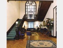 Atrio interno con scala monumentale in legno scuro e moquette verde che porta a un ballatoio con ringhiera decorata, davanti a una grande vetrata istoriata ad arco. Pavimento in parquet a spina di pesce, salottino con divani e poltrone, tappeti e grandi piante ornamentali lungo le pareti.