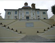 Facciata di un edificio storico con elementi barocchi, cupola e campanile, vista dal basso. Un’ampia scalinata centrale in pietra con rampe laterali e vasi di piante conduce a una terrazza davanti all’ingresso.