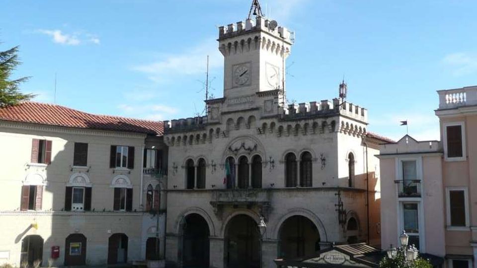 Edificio storico in pietra chiara con facciata ad archi e torre centrale merlata con orologio, affacciato su una piazza. Ai lati si vedono palazzi intonacati con tetti in tegole rosse e cielo sereno sullo sfondo.