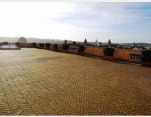 Ampia terrazza panoramica con pavimentazione in mattoni a spina di pesce e parapetto in muratura, fiancheggiata da vasi con siepi. Sullo sfondo si vede lo skyline urbano con cupole e campanili sotto un cielo chiaro.