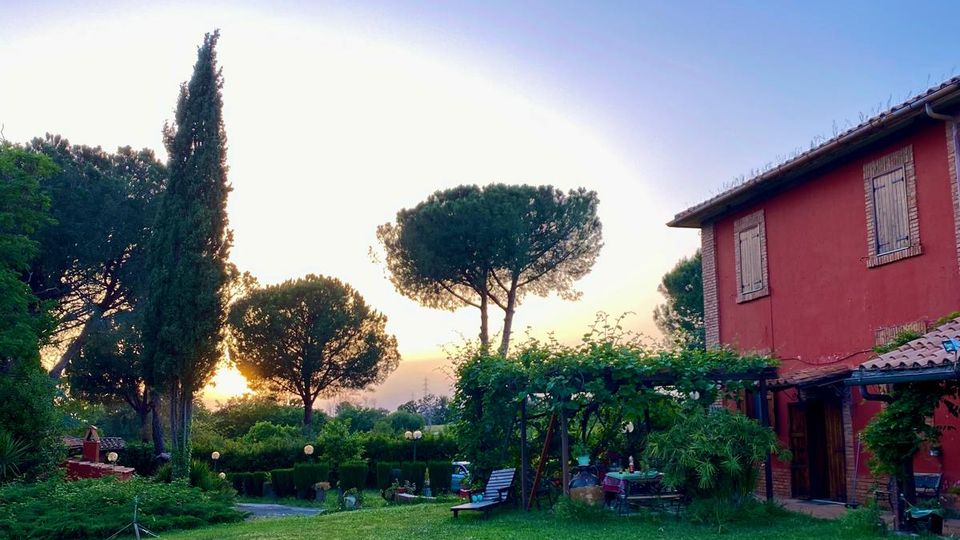 Ampio giardino con prato verde e alberi alti, con lampioni e sedute lungo un vialetto. Sulla destra si vede una casa di campagna a due piani con facciata rossa, persiane in legno e portico coperto da rampicanti.