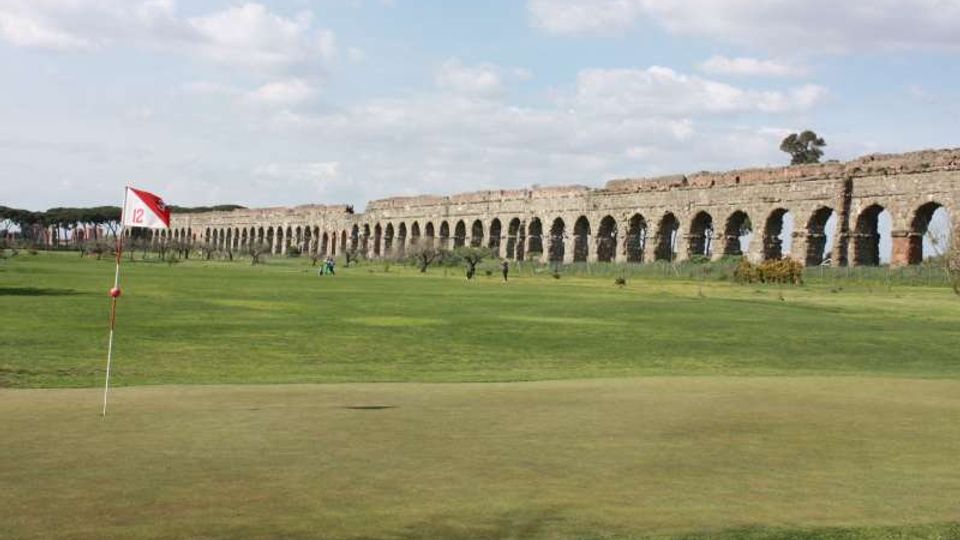 Campo da golf con bandierina sul green in primo piano, circondato da ampie aree erbose. Sullo sfondo si estende un lungo acquedotto in pietra con una serie di arcate sotto un cielo parzialmente nuvoloso.
