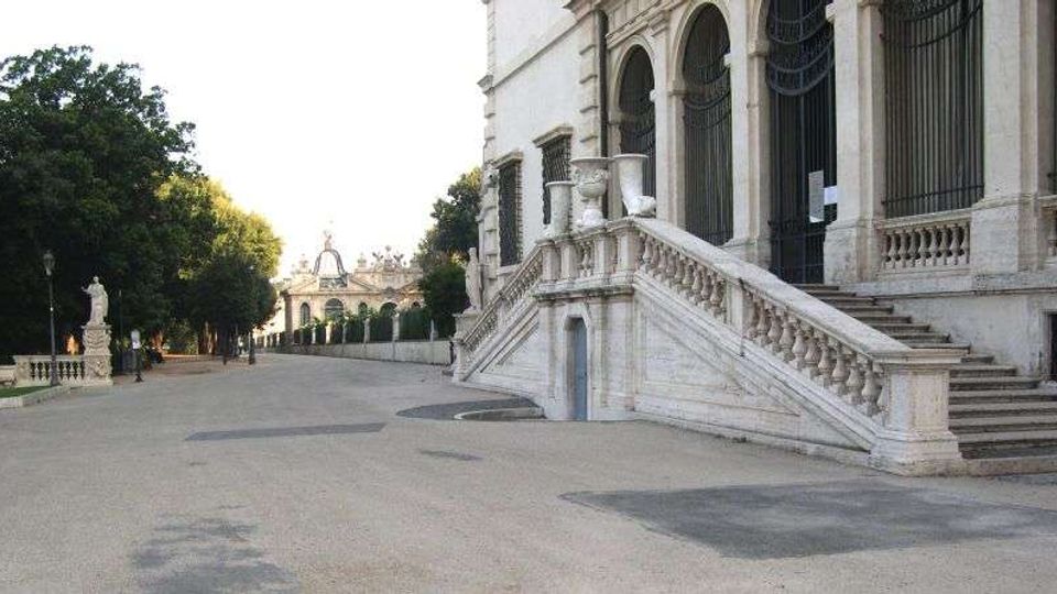 Ampio viale lastricato accanto a un edificio storico in pietra chiara, con grandi arcate e cancellate; una scalinata monumentale con balaustra conduce all’ingresso sopraelevato. A sinistra si vede un’area alberata con statue lungo il percorso e, sullo sfondo, strutture architettoniche simili a un portico.